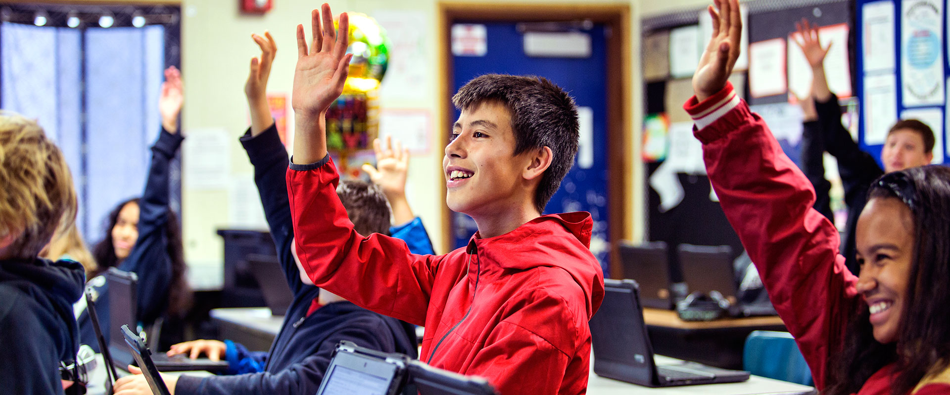 Elementary students in a computer lab raising their hands and smiling