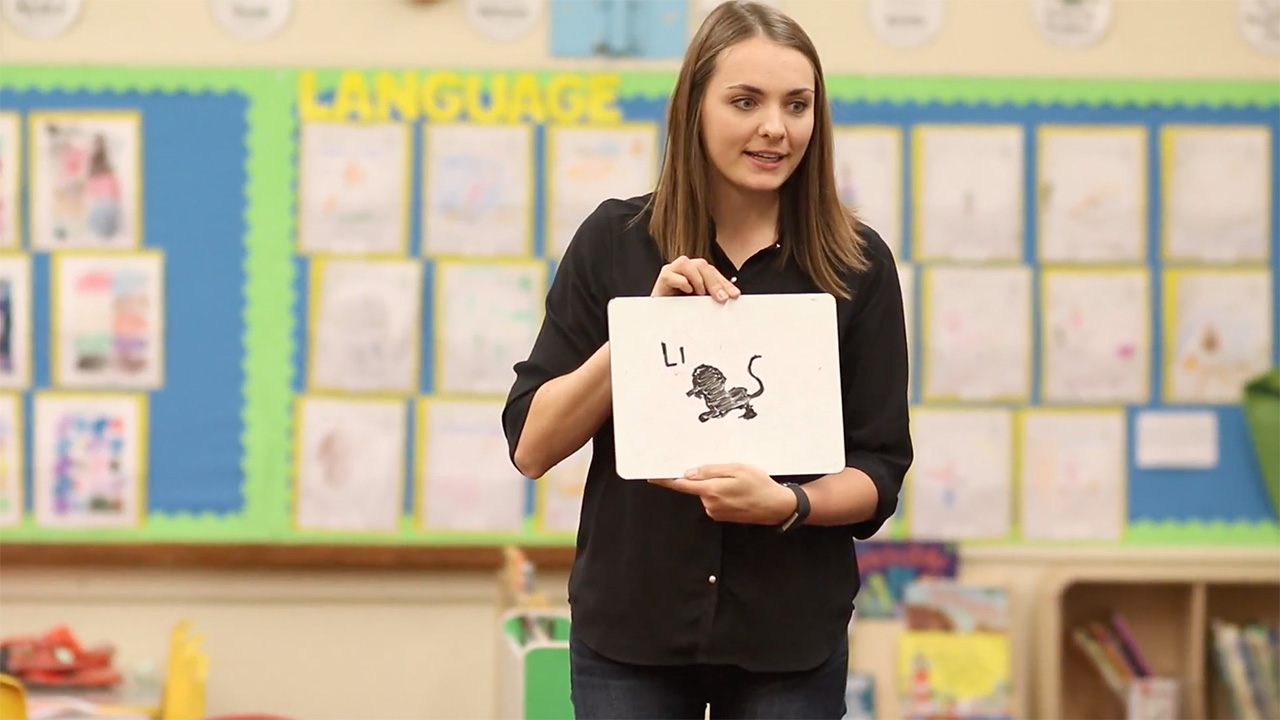 A teacher holding a small white board with an illustration of a lion asking for the proper spelling
