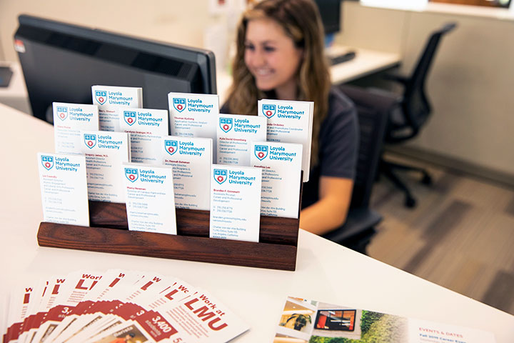 A student working behind a reception desk on campus with a rack of business cards on the counter