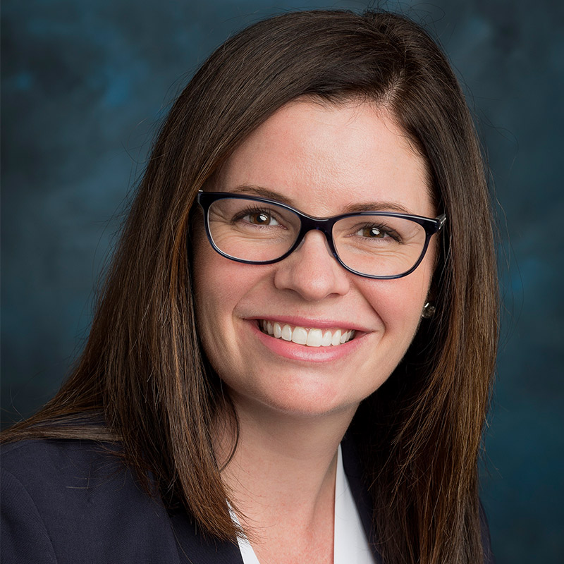 A woman with brown hair and black glasses smiling in front of a blue background