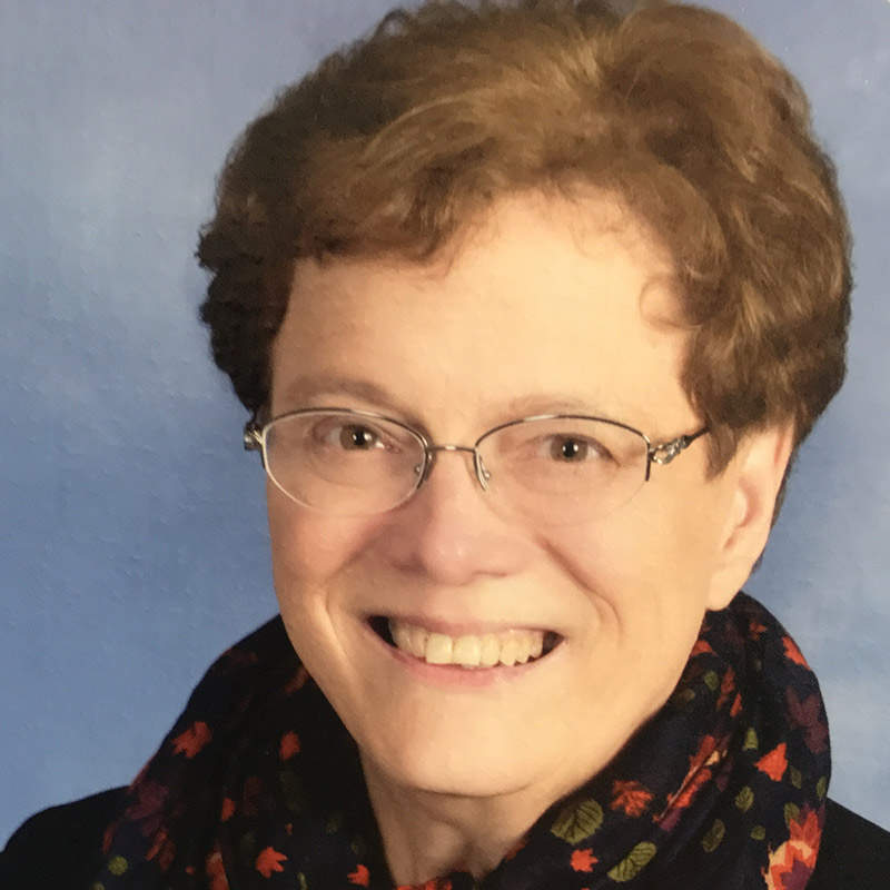 A Woman with short curly brown hair and glasses smiling in front of a light blue background