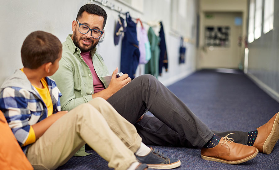 teacher sitting on the floor of a school hallway and talking to a student