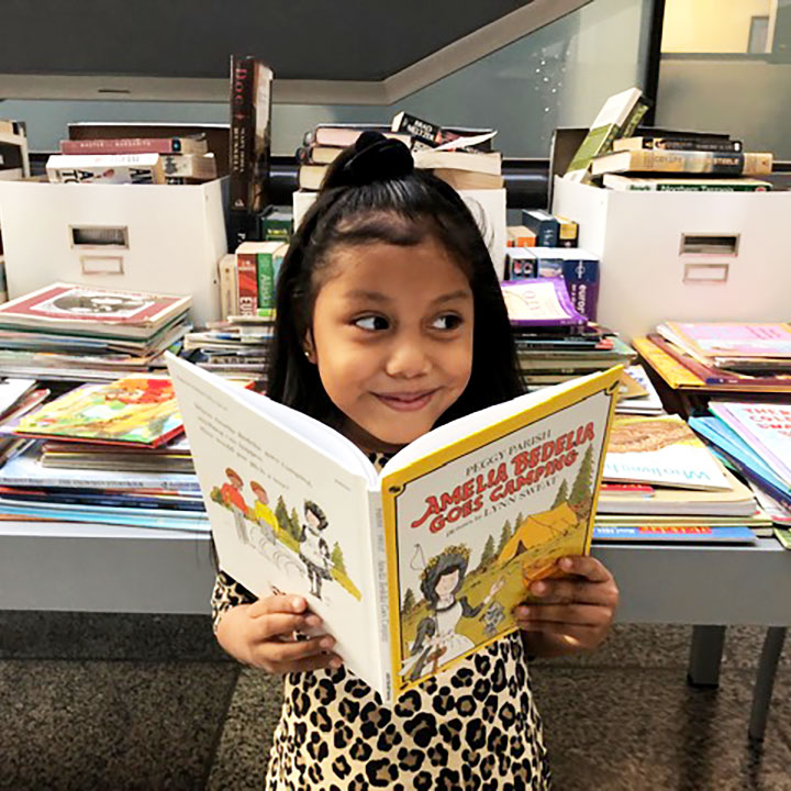 young girl student holding an open book in front of her and smiling while standing in front of a table of books