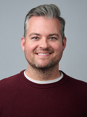 Korey Hlaudy Portrait, man with silver hair in maroon sweater, in front of a grey background