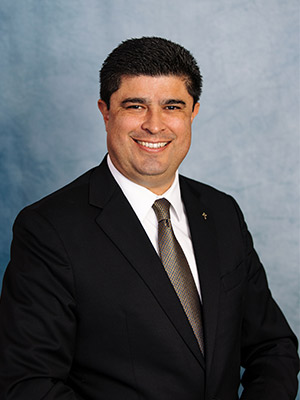 Paul Escala portrait dark haired man in black suit in front of a blue background