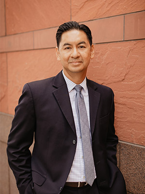 Manwith dark hair, dressed in black suit standing in front of a sandstone building